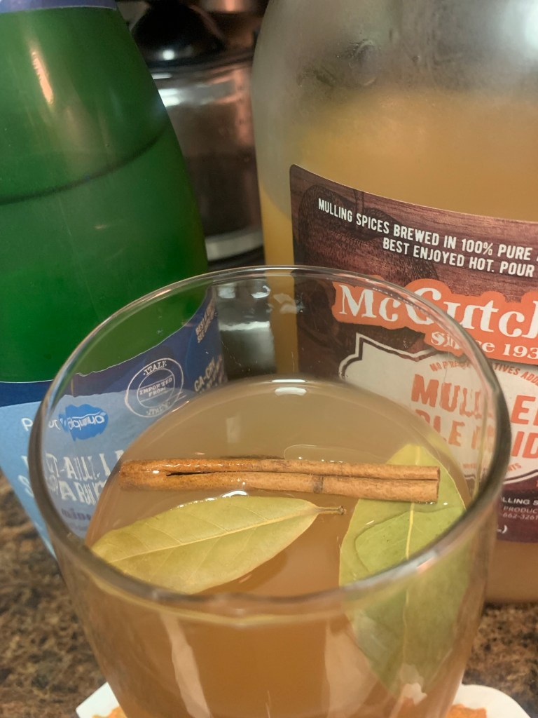 A glass of mulled apple cider, garnished with a cinnamon stick and bay leaves, is placed on a countertop next to a bottle of McCutcheon's mulling spices and a green bottle of sparkling water.