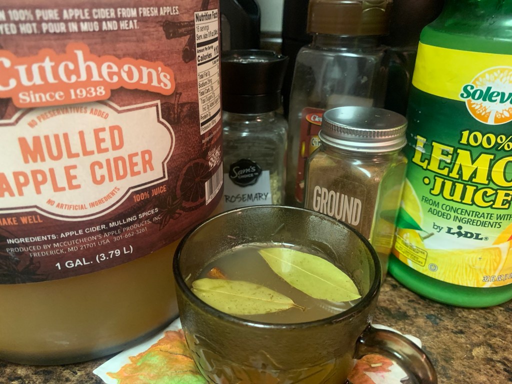 A glass mug filled with mulled apple cider, garnished with bay leaves, sitting on a countertop surrounded by various spices and lemon juice.